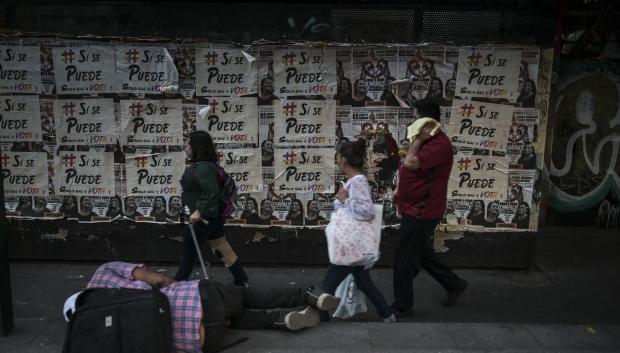 Un hombre duerme en una acera mientras la gente pasa junto a un muro con carteles de partidos de oposición y otros con la imagen de la candidata presidencial de México por el partido gobernante Morena, Claudia Sheinbaum