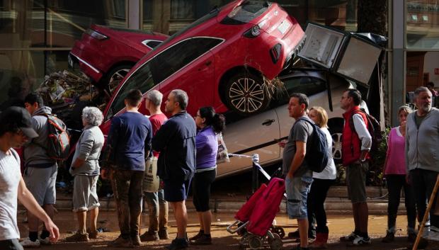 La gente hace cola para recibir suministros cerca de una montaña de coches destrozados por la DANA