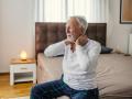 Senior adult man with gray hair and beard suffering from neck pain and stiffness, sitting on his bed in pajamas, feeling discomfort in the morning at home