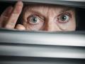 A senior woman peeps fearfully through venetian blinds