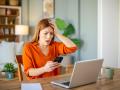 Frustrated worried businesswoman sitting at home office in front of laptop, looking at mobile phone, holding head.