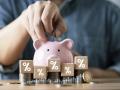 Close-up of a person putting a coin into a pink piggy bank with stacked coins and percentage blocks representing saving money financial growth investment and interest rate concept.