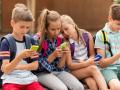 primary education, friendship, childhood, technology and people concept - group of happy elementary school students with smartphones and backpacks sitting on bench outdoors