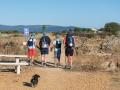 A family is walking the Camino de Santiago with their small pet dog. They are wearing similar hats, and the bags they are carrying have scallop shells attached to them to prove they are pilgrims.