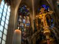 Close-up of ornate golden crucifix in a church interior, with blurred stained glass windows and vaulted ceiling in the background.