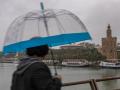 Un hombre se protege con paraguas y chubasquero de la lluvia y el viento mientras camina frente a la Torre del Oro en Sevilla