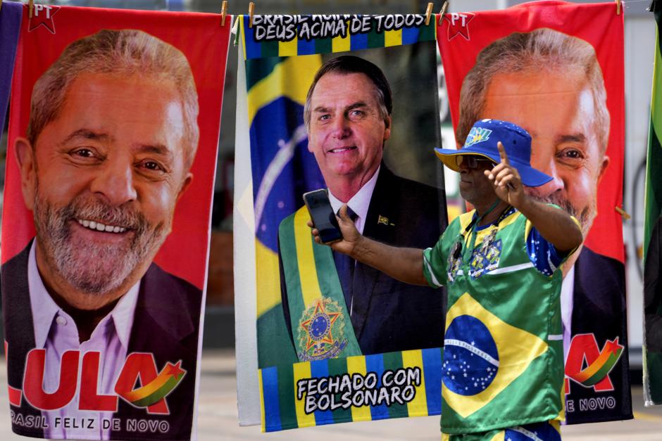 A demonstrator dressed in the colors of the Brazilian flag performs in front of a street vendor's towels for sale featuring Brazilian presidential candidates, current President Jair Bolsonaro, center, and former President Luiz Inacio Lula da Silva in Brasilia, Brazil, Tuesday, Sept. 27, 2022. Brazilians head to polls on Oct. 2 to elect a president, vice president, governors and senators. (AP Photo/Eraldo Peres)