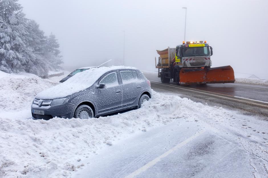 Un coche cubierto de nieve y una máquina quitanieves en la carretera de acceso al Puerto de Navacerrada