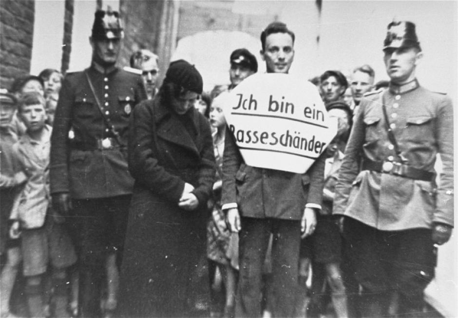 Flanqueado por policías alemanes, el joven lleva un cartel que dice, "Soy un corruptor de la raza"