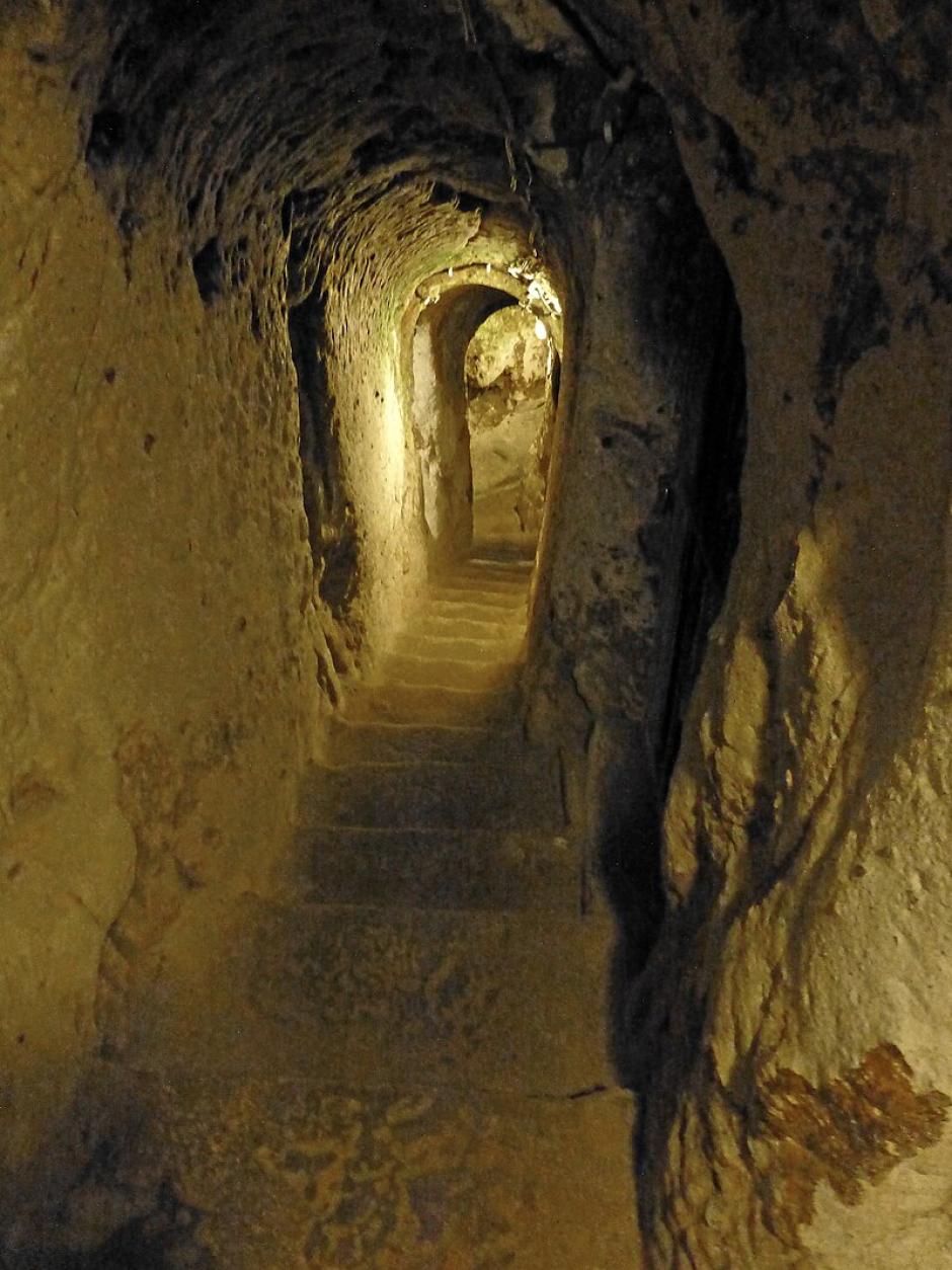 Pero debajo de la superficie desmoronada de Capadocia, una maravilla de proporciones igualmente gigantescas estuvo escondida durante siglos; una ciudad subterránea que podía ocultar el paradero de hasta 20.000 habitantes durante meses seguidos.
85 metros bajo tierra
La antigua ciudad de Elengubu, conocida hoy como Derinkuyu, se encuentra a más de 85 metros bajo la superficie de la Tierra, abarcando 18 niveles de túneles.
Es la ciudad subterránea excavada más grande del mundo y estuvo en uso casi constante durante miles de años, cambiando de manos de los frigios a los persas a los cristianos de la era bizantina.
Finalmente fue abandonada en la década de 1920 por los griegos de Capadocia cuando enfrentaron la derrota durante la guerra greco-turca y huyeron abruptamente en masa a Grecia.
Una habitación dentro de la ciudad subterráneaFUENTE DE LA IMAGEN,GETTY IMAGES
No solo sus habitaciones con forma de cueva se extienden por cientos de kilómetros, sino que se cree que las más de 200 pequeñas ciudades subterráneas separadas que también se han descubierto en la región pueden estar conectadas a estos túneles, creando una red subterránea masiva.
Según mi guía, Suleman, Derinkuyu solo fue "redescubierto" en 1963 por un lugareño anónimo al que constantemente se le perdían sus pollos.
Mientras renovaba su casa, las aves de corral desaparecían en una pequeña grieta creada durante la remodelación y nunca más se las volvía a ver.
Tras una investigación más detallada y algunas excavaciones, el turco descubrió un pasadizo oscuro.
Fue la primera de más de 600 entradas encontradas dentro de casas particulares que conducen a la ciudad subterránea de Derinkuyu.
Civilización escondida
La excavación comenzó de inmediato, revelando una red enredada de viviendas subterráneas, almacenamiento de alimentos secos, establos de ganado, escuelas, bodegas e incluso una capilla.
Era toda una civilización escondida a salvo bajo tierra.
La ciudad cueva pronto fue visitada por miles de los turistas menos claustrofóbicos de Turquía y, en 1985, la región se agregó a la lista del Patrimonio Mundial de la Unesco.
Vista de túneles bajo tierraFUENTE DE LA IMAGEN,GETTY IMAGES
Pie de foto,Derinkuyu se compone de 18 niveles de túneles.
La fecha exacta de construcción de la ciudad sigue siendo discutida, pero "Anabasis", escrita por Jenofonte de Atenas alrededor del 370 a.C., es la obra escrita más antigua que parece hacer referencia a Derinkuyu.
En el libro, menciona a la gente de Anatolia, en o cerca de la región de Capadocia, que vive bajo tierra en casas excavadas en lugar de en las más populares cuevas cavadas en los acantilados también bastante conocidas en la zona.