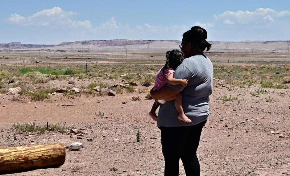 A utility pole is seen on the ground, with power poles offering electricity in the distance lining Highway 89, as Navajo Nation resident Gilberta Cortes, 42, who uses a generator for electricity, carries her child outside her home in Cameron, Arizona, on July 9, 2025 as she waits for electricity from the power grid to reach her residence. Initiatives like "Light Up Navajo," a project launched by the semi-autonomous government of the reservation in 2019, are connecting homes to the power grid with help from volunteer utility crews and federal funding but challenges remain due to the dispersed nature of the communities and vastness of the reservation. (Photo by Frederic J. BROWN / AFP)
