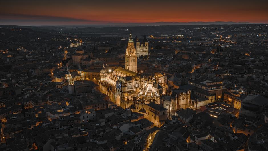 Aerial night view of Toledo Cathedral illuminated above the historic old town of Toledo in central Spain. The Gothic cathedral stands out against a dense network of medieval streets and tiled rooftops, with warm city lights glowing beneath a dark red evening sky, highlighting the architectural and cultural heritage of this UNESCO World Heritage city. Castilla–La Mancha, central Spain, Europe.