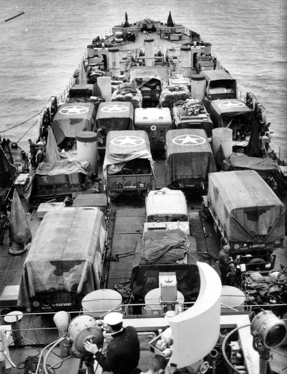 Vehículos ingleses en la cubierta superior de un LST Estadounidense. Muchos vehículos están pintados y cubiertos con lonas de camuflaje. La División Tyne and Tees (TT) desembarcó en Gold Beach el 6 de junio.
